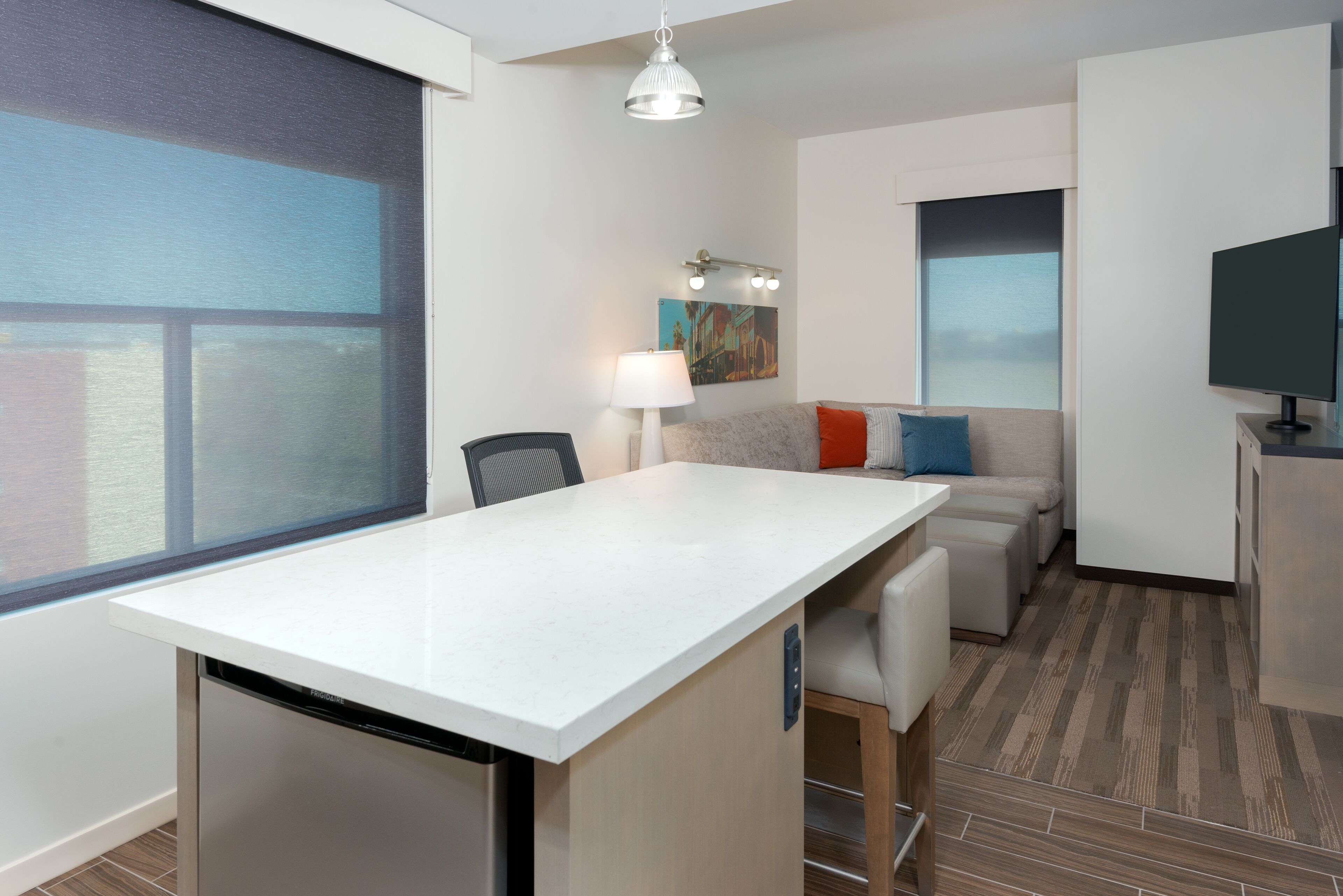A modern hotel room with a large window, a dining area, a sofa, a television, and a lamp.
