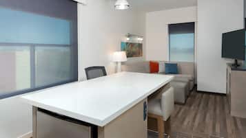 A modern hotel room with a large window, a dining area, a sofa, a television, and a lamp.