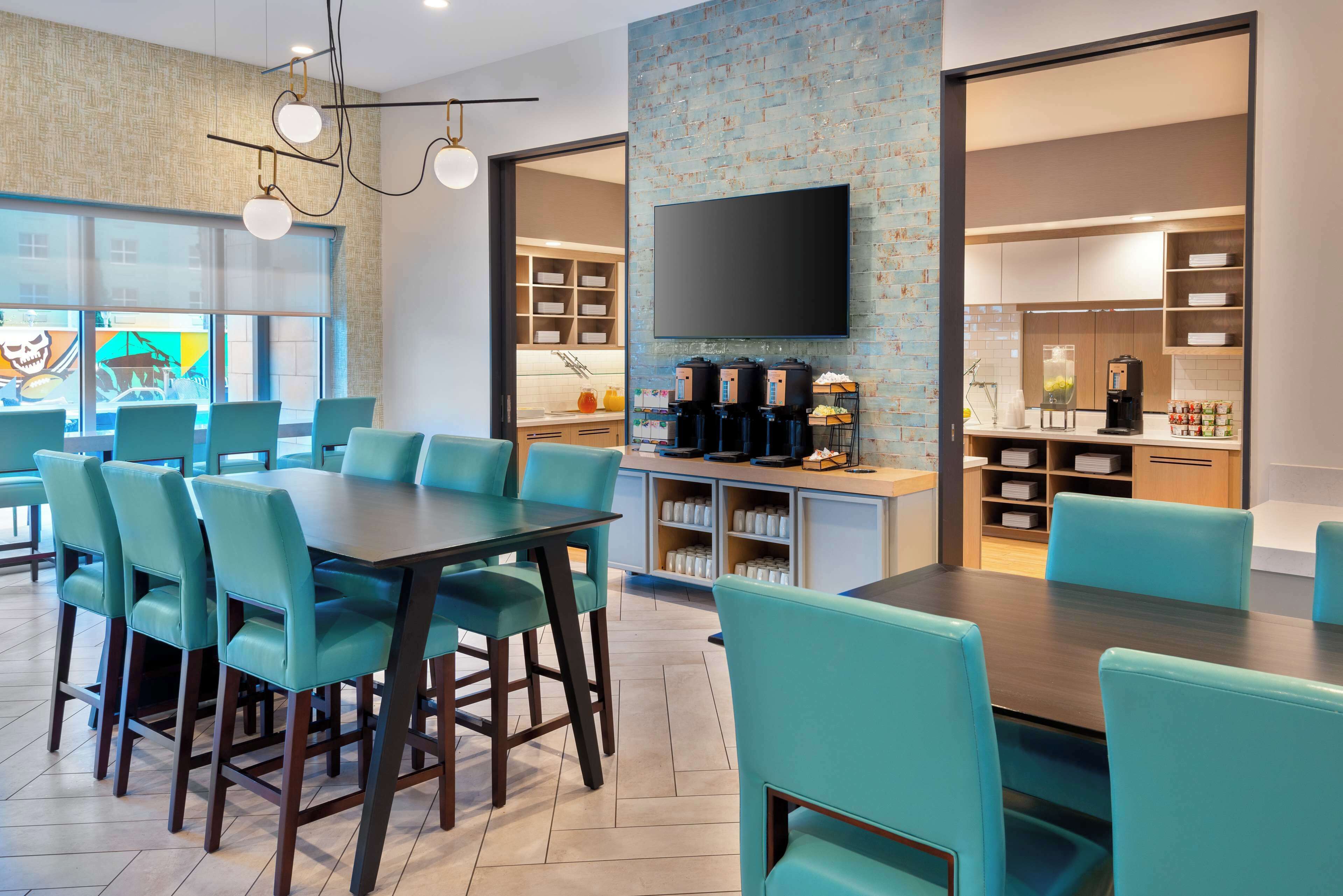 A modern dining area with teal chairs and a dark wooden table, a kitchen with a flat-screen TV, and a visible coffee machine.