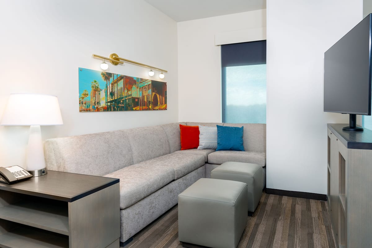 A modern hotel room with a sofa, ottoman, bedside table with a lamp, a painting on the wall, and a TV.