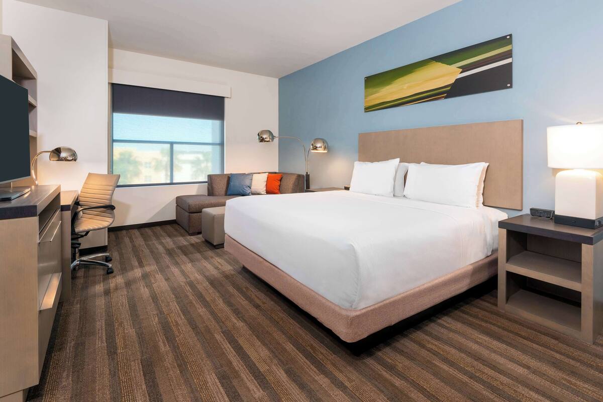 A hotel room with a large bed, a desk with a computer, a chair, and a window with blinds.