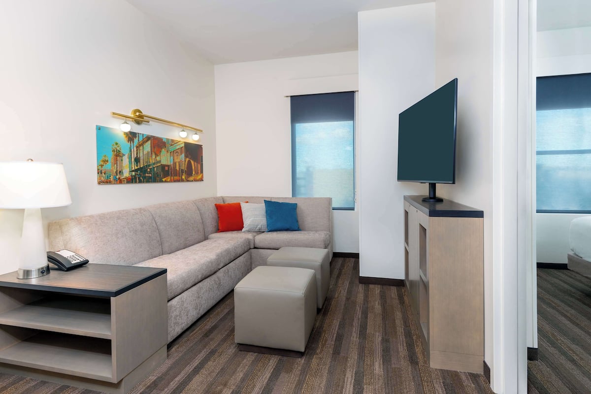 A modern hotel room with a sectional sofa, a small table with a lamp, a flat-screen TV, and a painting on the wall.
