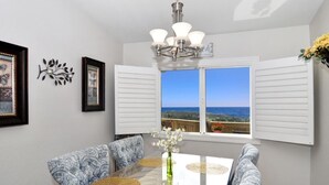 A dining area with a glass table, patterned chairs, a chandelier, and a view of the ocean through large windows. - Panoramic Ocean View-No Stairs-King Bed-Walkin shower, 24hr Manned Sec. Gates (Dana Point)