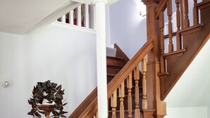 A wooden staircase with a white column, a decorative urn, and a wicker chair. - Historic Vermont Farmhouse on 12 Acres, Quaint Vermont Experience. (Andover)