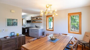 Une cuisine moderne dotée d’un îlot central, d’un coin repas avec une table en bois et d’une vue sur les arbres par une fenêtre.
