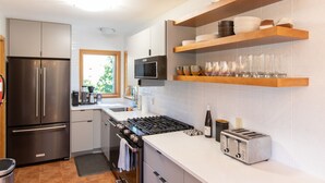 Une cuisine moderne équipée d’un réfrigérateur, d’un four et d’une cuisinière en acier inoxydable, de tablettes en bois au-dessus du plan de travail et d’une fenêtre donnant sur un espace verdoyant.