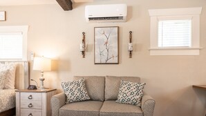 Een gezellige woonkamer met een grijze bank, een bijpassend poefje en een kamerkleed met een patroon. Er staat een klein wit nachtkastje met lades en een lamp. Een ingelijste foto hangt aan de muur boven de bank.