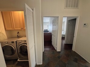 A laundry area with a washing machine and dryer, a wooden cabinet, and a hallway leading to a bedroom with a bed and a nightstand.