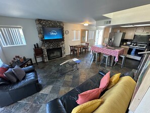 A living room with a stone fireplace, a flat-screen TV mounted on the wall, a dining area with a table and chairs, and a kitchen with modern appliances.
