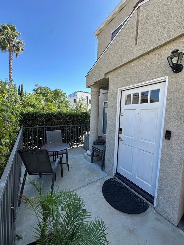 A residential building with a white door, a small patio with a table and chairs, a black metal railing, and a palm tree in the background.