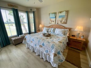 Une chambre à coucher avec un grand lit, deux tables de chevet, un banc et une vue sur l’extérieur grâce à de grandes fenêtres.