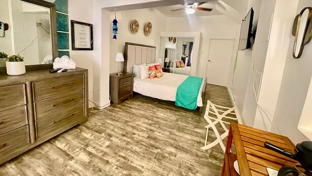 A bedroom with a bed, bedside table, dresser, mirror, ceiling fan, and a wooden chair. - Casa Sevilla, Coqui Studio (San Juan)