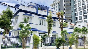 A two-story building with a white facade and blue accents, surrounded by a black fence and potted plants. - Casa Sevilla, Coqui Studio (San Juan)