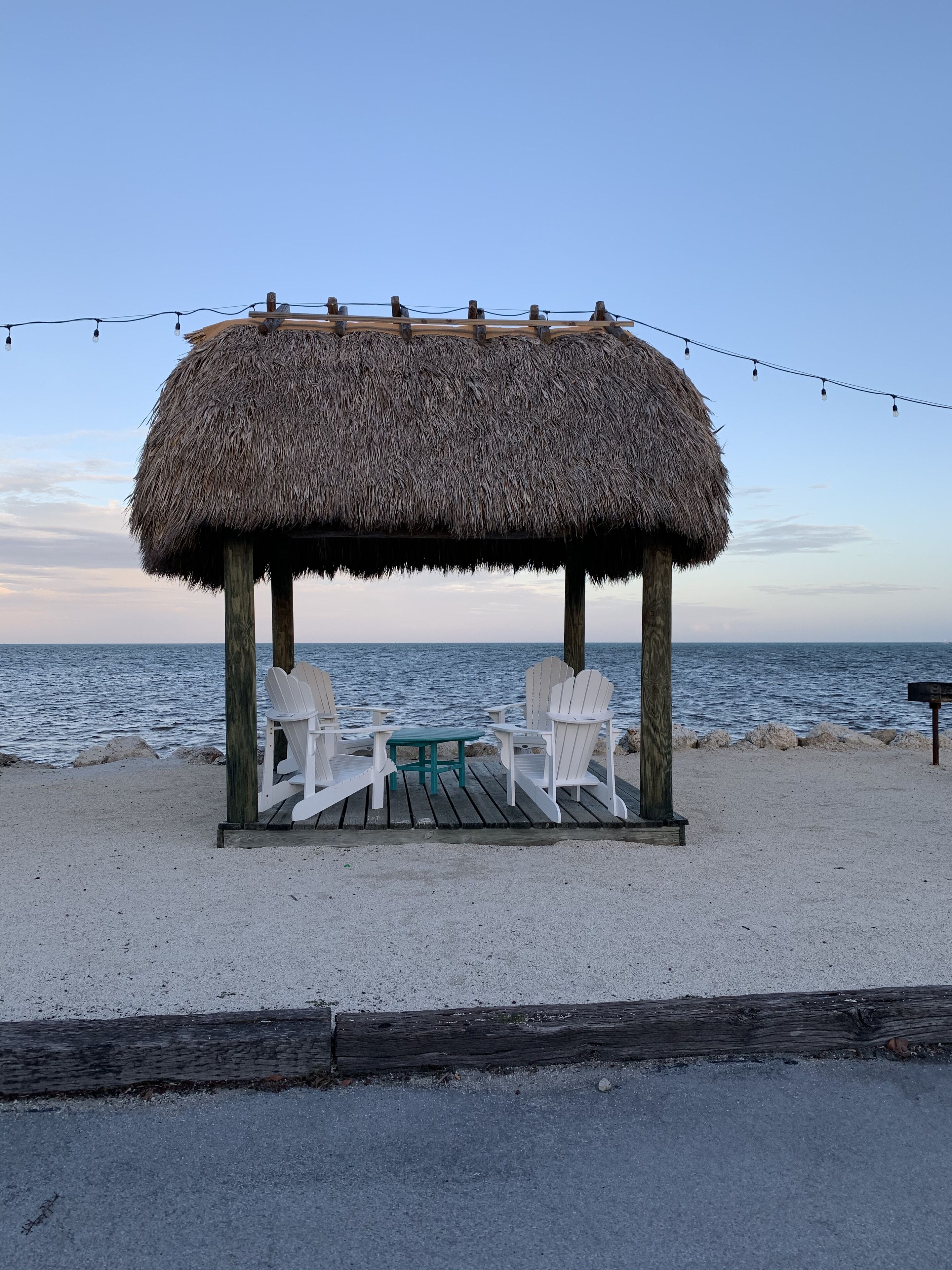 A beachside gazebo with a thatched roof, white Adirondack chairs, and a small table.