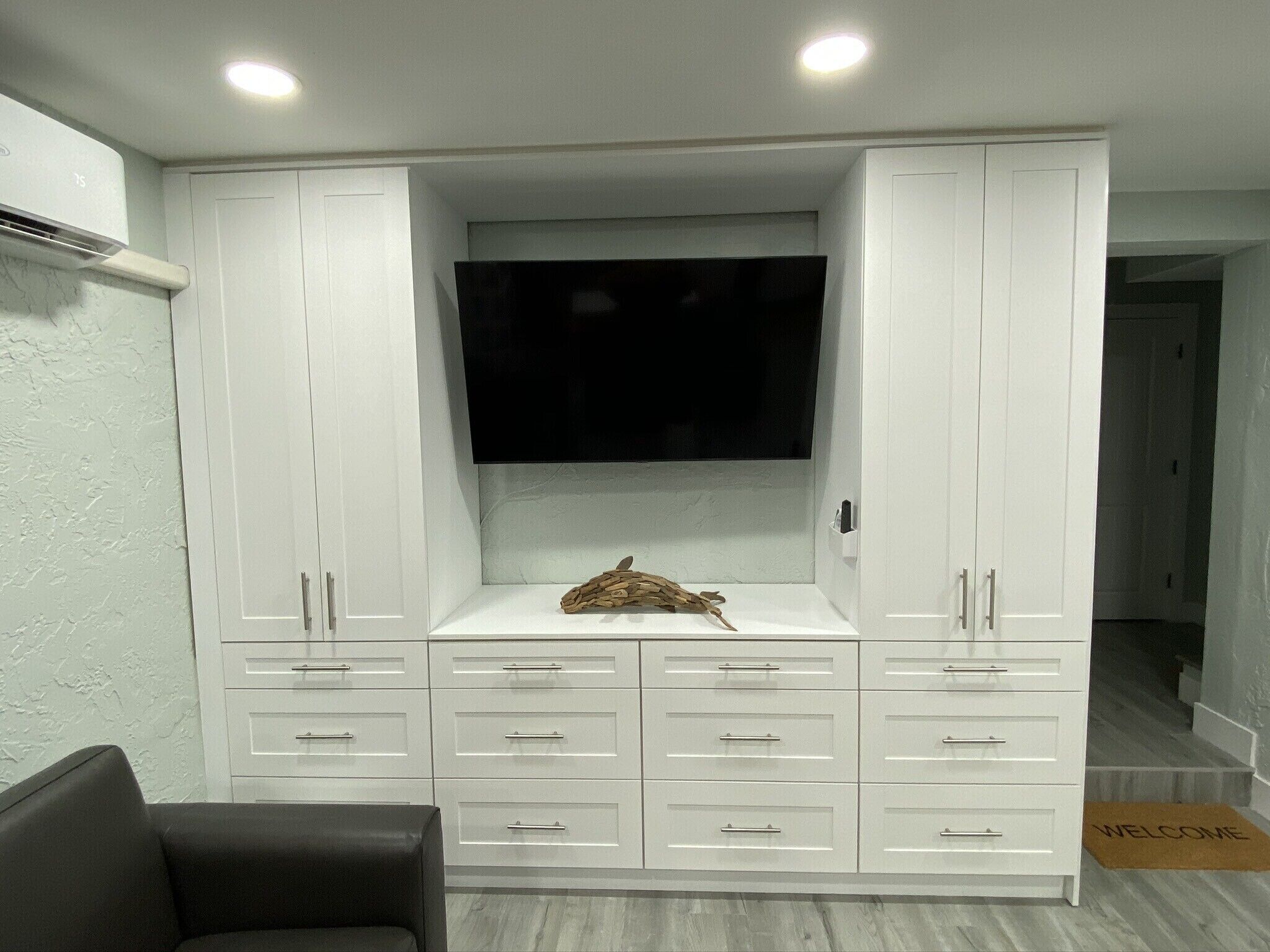 Een moderne woonkamer met een flatscreen televisie bevestigd aan een wit kastje, een houten decoratie op het onderste plankje, en een 'WELCOME' mat bij de ingang.