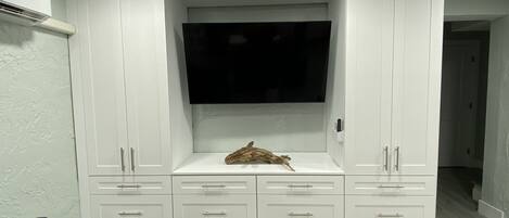 Een moderne woonkamer met een flatscreen televisie bevestigd aan een wit kastje, een houten decoratie op het onderste plankje, en een 'WELCOME' mat bij de ingang.