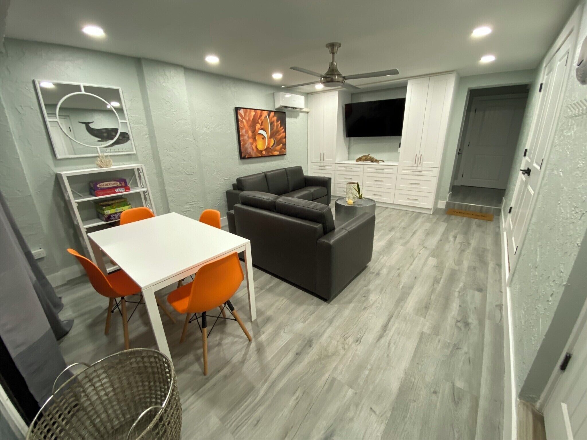 A modern living room with a grey sofa, a white dining table with orange chairs, a ceiling fan, and a TV on the wall.