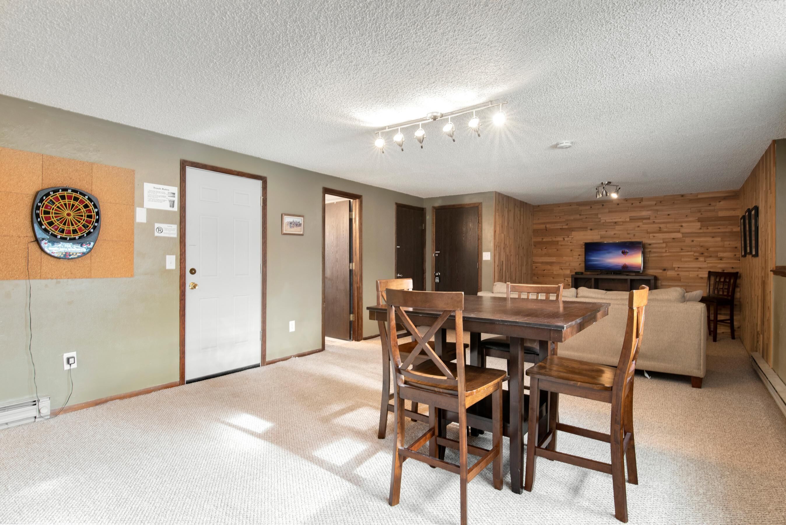 A spacious living area with a dining table and chairs, a television, and a dartboard on the wall.
