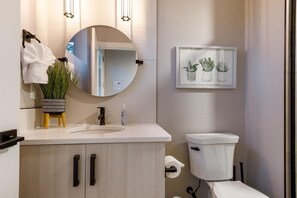 A modern bathroom with a round mirror, a sink, a toilet, and a framed plant artwork on the wall.