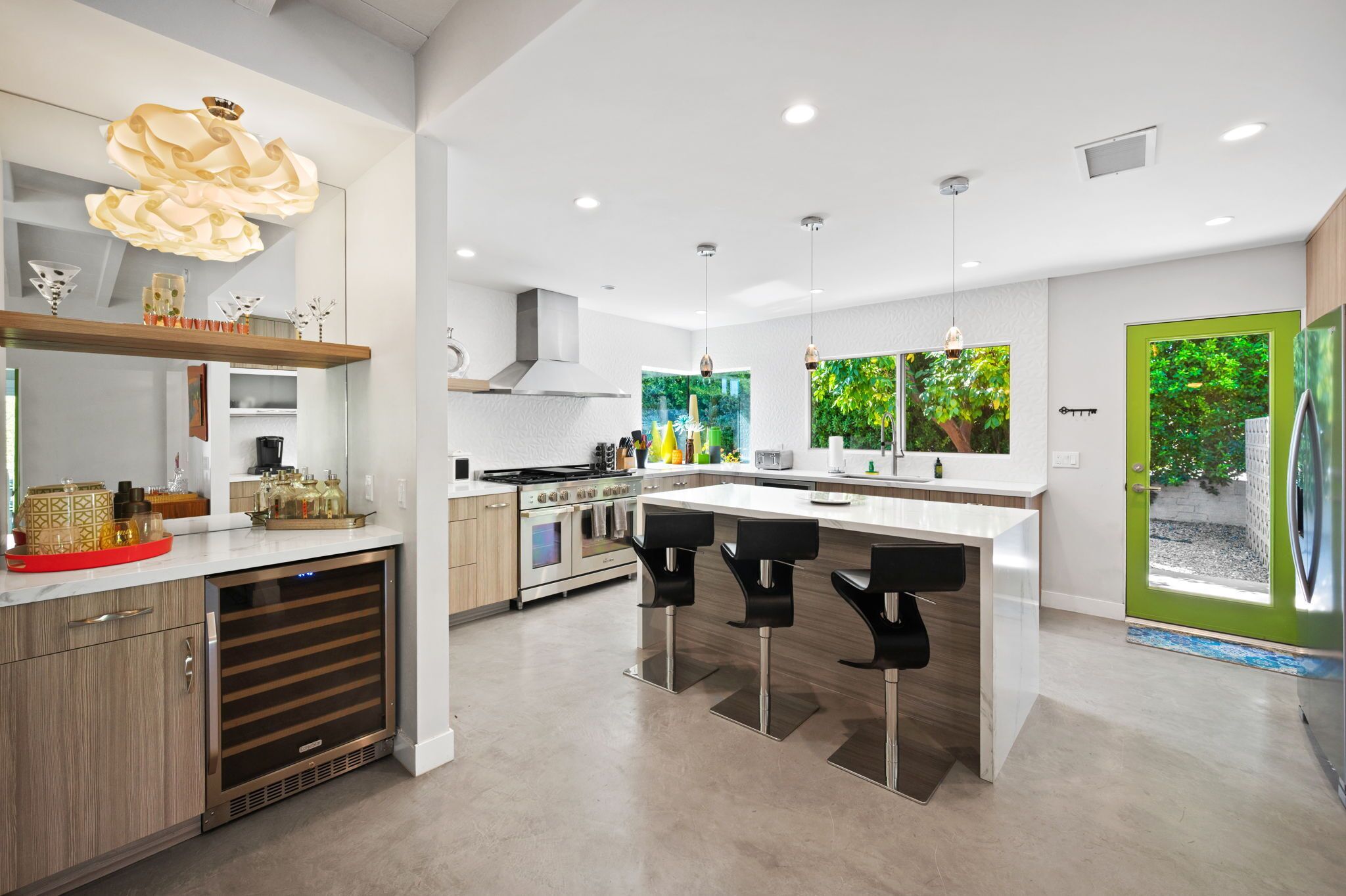 A modern kitchen with a central island, bar stools, a built-in oven, and a large window with a view of greenery.