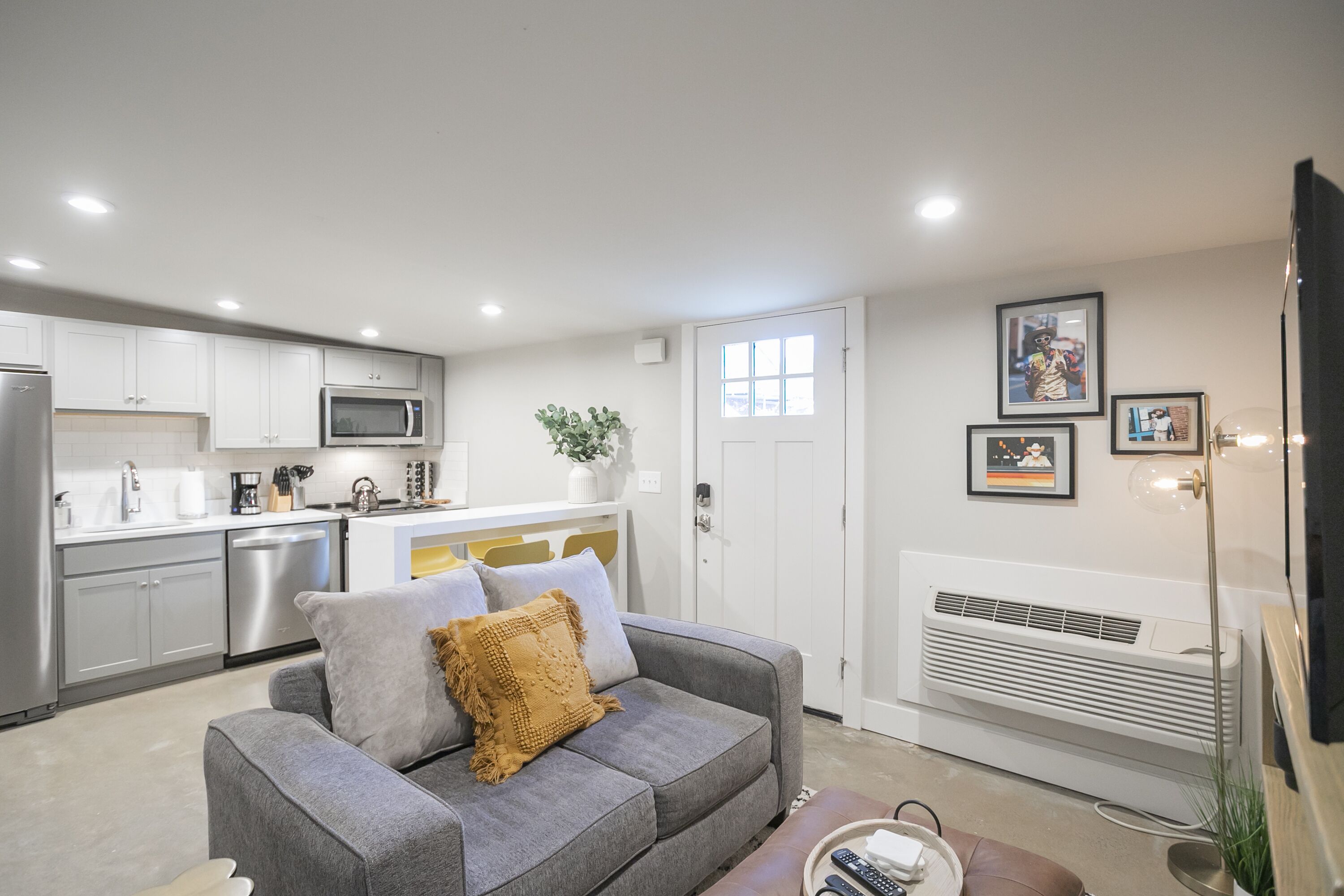 A modern kitchen with white cabinets, a microwave, and a dishwasher. There is a gray sofa with cushions and a small table with a remote control.