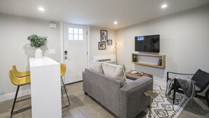 A modern living room with a grey sofa, yellow chairs, a small table with a plant, a TV on a shelf, and a door. - Serene Guest House - Foodie Oasis - Walk to Folk, Audrey, Red-Headed Stranger! (Nashville)