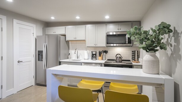 A modern kitchen with white cabinets, a stainless steel refrigerator, a microwave, and a central island with yellow chairs. - Serene Guest House - Foodie Oasis - Walk to Folk, Audrey, Red-Headed Stranger! (Nashville)