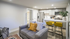 A modern living room with a grey sofa, a yellow throw pillow, and a coffee table. There is a kitchen area with white cabinets, a microwave, and a sink. - Serene Guest House - Foodie Oasis - Walk to Folk, Audrey, Red-Headed Stranger! (Nashville)