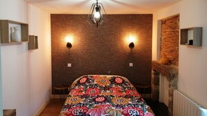 Ein Schlafzimmer mit einer Wand im Backsteinmuster, zwei Wandleuchten, einem Deckenventilator, einem Bett mit bunter Blumenbettdecke und einem kleinen Tisch mit Stühlen.