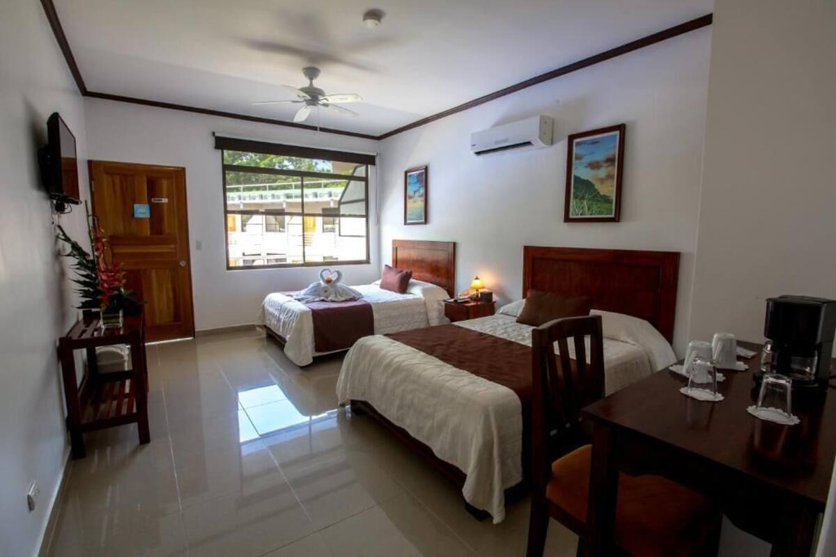 A hotel room with two beds, a desk with a chair, a ceiling fan, and a window with a view of buildings outside.