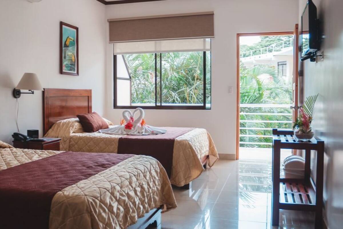 A hotel room with two beds, a large window with a view of greenery, a bedside table with a lamp, and a small table with a vase.