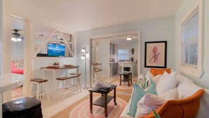 A living room with a TV, white furniture, a sofa with colorful cushions, and a kitchen in the background.