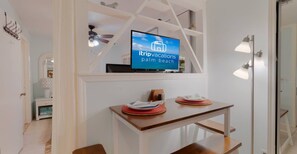 Una sala de estar moderna con una televisión mostrando 'itripvacations palm beach', una mesa de comedor con platos y un ventilador de techo.