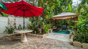 A patio with a stone table, a red umbrella, and a pool area surrounded by lush greenery.