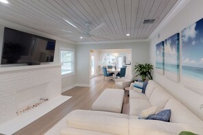 A modern living room with a white brick fireplace, a flat-screen TV mounted above it, a large white sofa with blue cushions, and a dining area in the background.