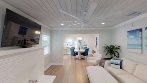 A modern living room with a flat-screen TV, a ceiling fan, and a dining area with blue chairs. - The Lewis Properties (West Palm Beach)