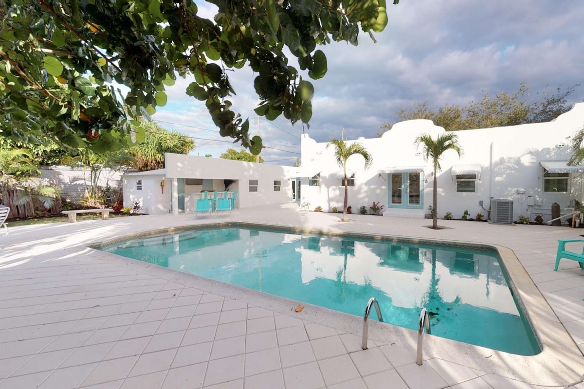 Una piscina con bordo piastrellato, un piccolo patio con sedie e un edificio bianco con una porta e finestre.