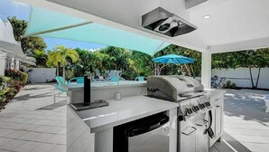 An outdoor kitchen with a grill, a sink, and a built-in oven. There are umbrellas providing shade, and a pool in the background. - The Lewis Properties (West Palm Beach)