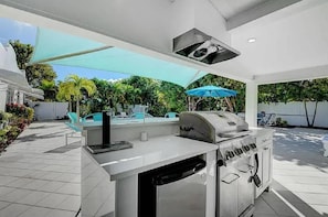 Cucina esterna con barbecue, lavello e forno integrato. Sono presenti ombrelloni per creare ombra, e una piscina sullo sfondo.