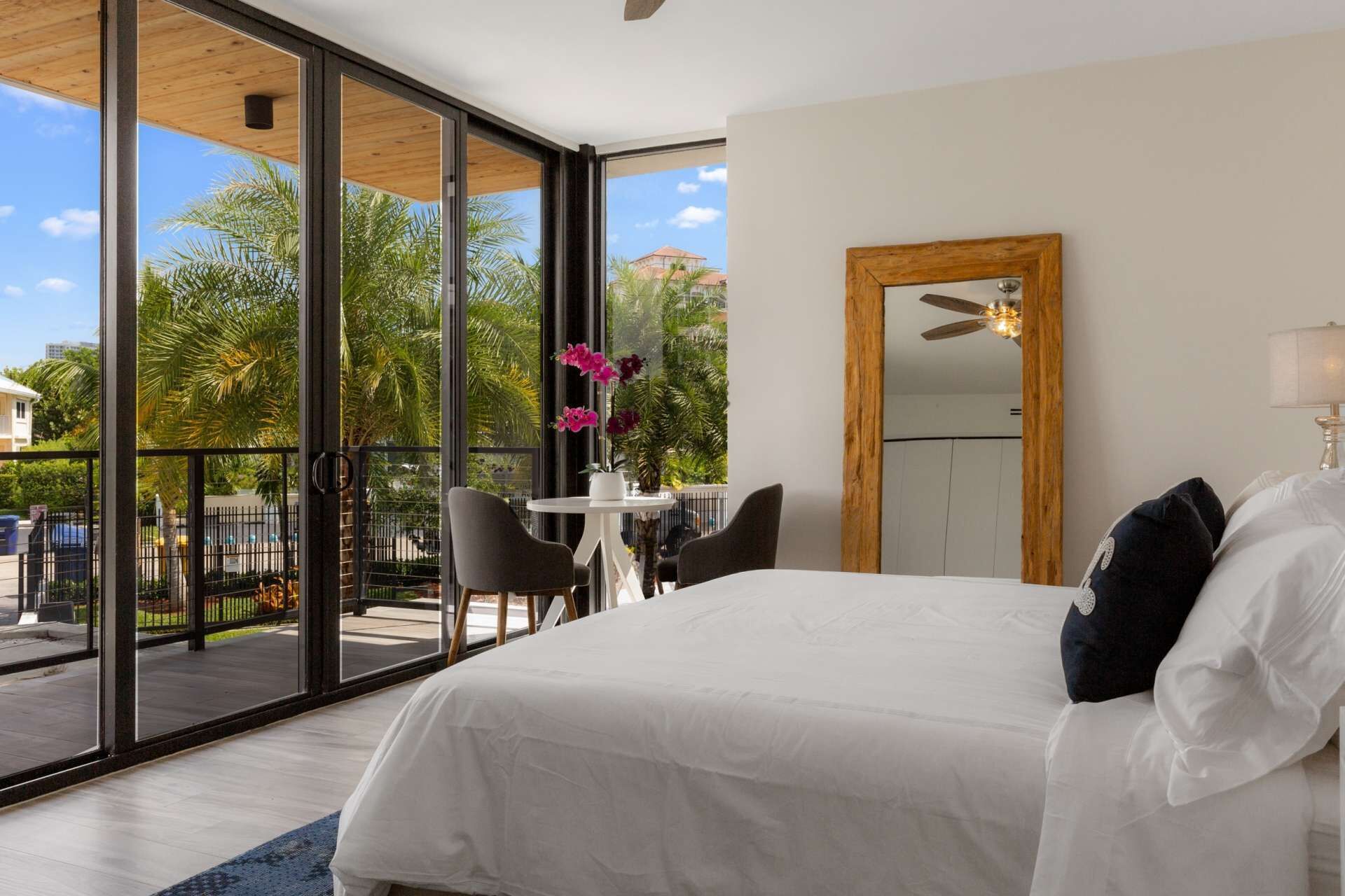 Ein modernes Hotelzimmer mit einem großen Bett, einem Esstisch mit Stühlen, einem Balkon mit Glastüren und Blick auf Palmen und Gebäude.