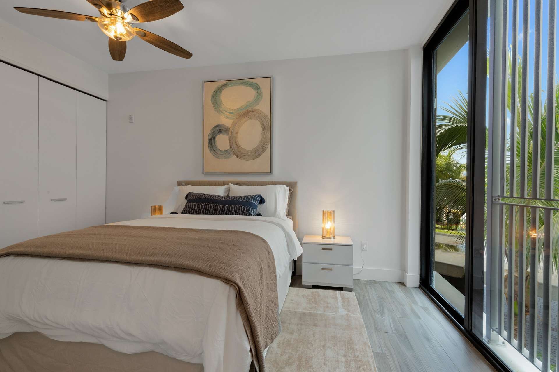A bedroom with a bed, a ceiling fan, a bedside table with a lamp, a large abstract painting, and a view of palm trees through a glass door.