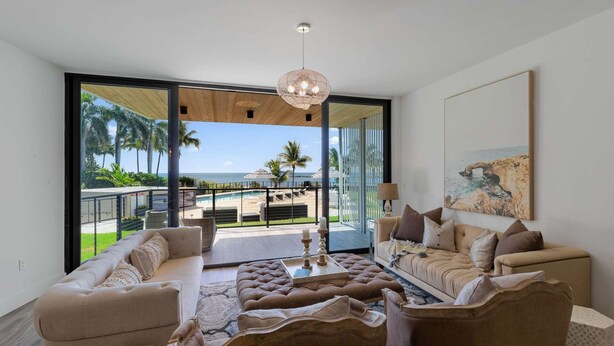 A spacious living room with a large sliding glass door leading to a balcony with a pool and palm trees. - NEW Luxurious Condo/inlet & Ocean Views 106 Inlet Way Unit 103 - Palm Beach Shores (Singer Island)