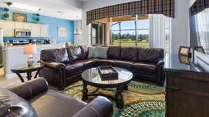 A living room with a leather sectional sofa, a glass coffee table, and a view of a golf course through large windows. - Welcome to The Windsor Dream! (Kissimmee)