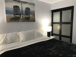 Een bed met witte lakens, een zwart geweven sprei, en een schilderij van twee stoelen op het strand.