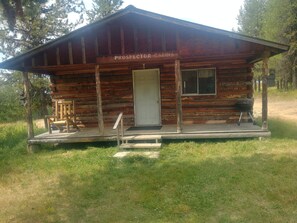 A rustic cabin labeled "Prospector Cabins" with a porch, a door, and a grill outside. - Prospector Hitching post (Elk City)