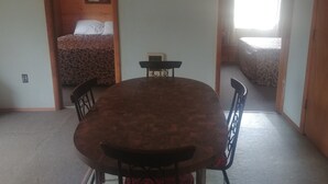 A room with a wooden dining table, two chairs, a bed with a patterned bedspread, and a window with curtains.