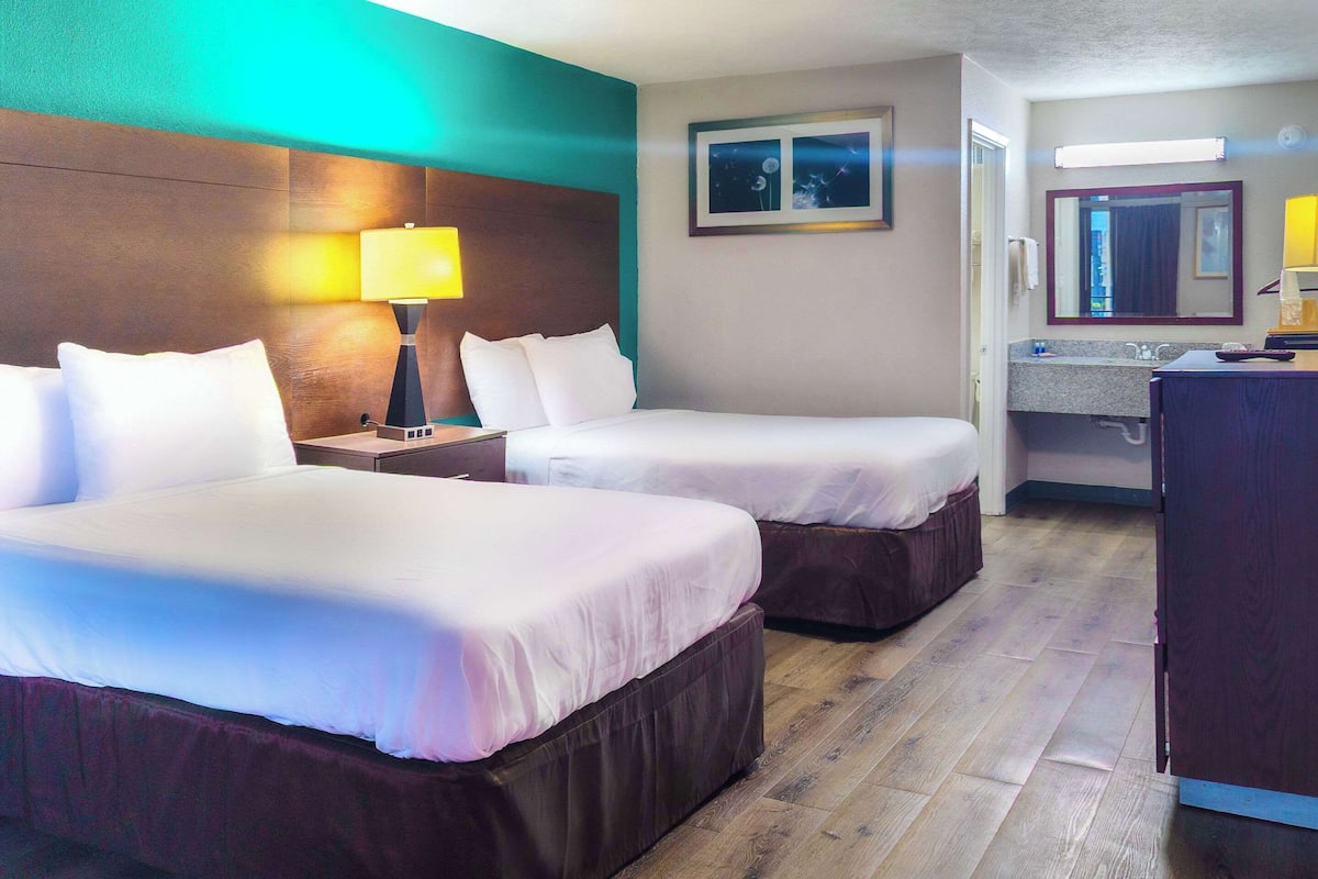 Une chambre d’hôtel avec deux lits, une table de chevet avec une lampe, une salle de bain avec un miroir et un plancher en bois.