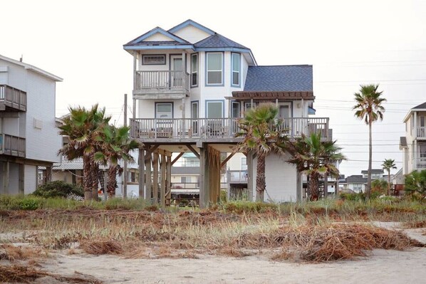 A two-story house with a balcony, surrounded by palm trees and a sandy beach. - Gorgeous Beachfront Home with Modern Finishes (Galveston)