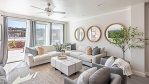 A modern living room with a sectional sofa, a coffee table, and a large window with curtains. There are two round mirrors on the wall and a ceiling fan. - Poolside Escape At The Ledges (St. George)