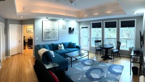 A modern living room with a blue sofa, a glass coffee table, and a dining area with a round table and chairs. There are large windows with blinds, a ceiling fan, and artwork on the walls. - Zen themed private retreat in NW Portland. (Portland)